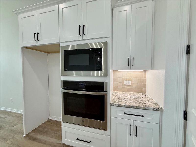 Built-in stainless steel microwave and oven, white shaker-style cabinetry with matte black hardware, under-cabinet lighting, natural stone backsplash, and granite countertops