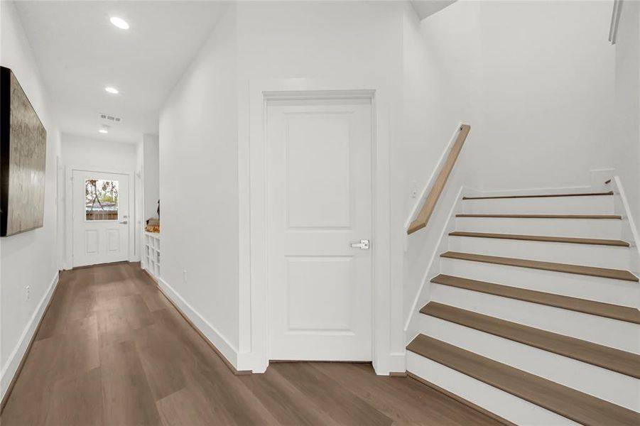 Well-lit hallway with wooden flooring, a staircase, and a white door. The open space is bright, leading to Two downstairs bedrooms and a patio area.