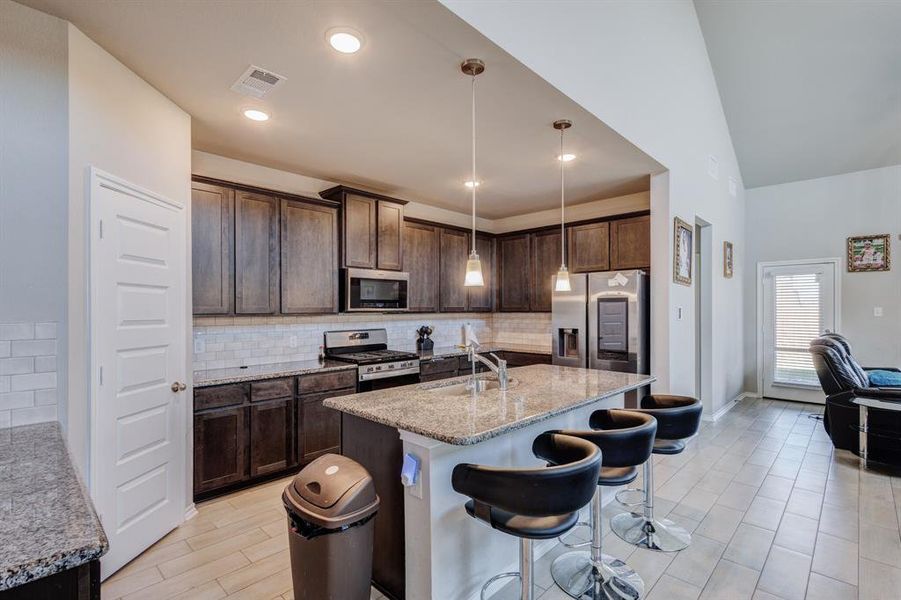 Kitchen featuring decorative backsplash, dark brown cabinetry, decorative light fixtures, a breakfast bar area, and light stone countertops Kitchen featuring decorative backsplash, dark brown cabinetry, decorative light fixtures, a breakfast bar area, and light stone countertops
