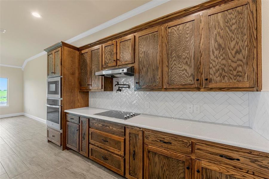 Kitchen featuring under cabinet range hood, crown molding, black electric stovetop, decorative backsplash, and light countertops