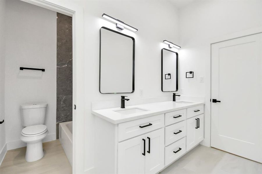 Secondary bath features tile flooring, bath/shower combo with tile surround, light colored cabinets, beautiful light countertop, vanity lights, and sleek, dark modern finishes.