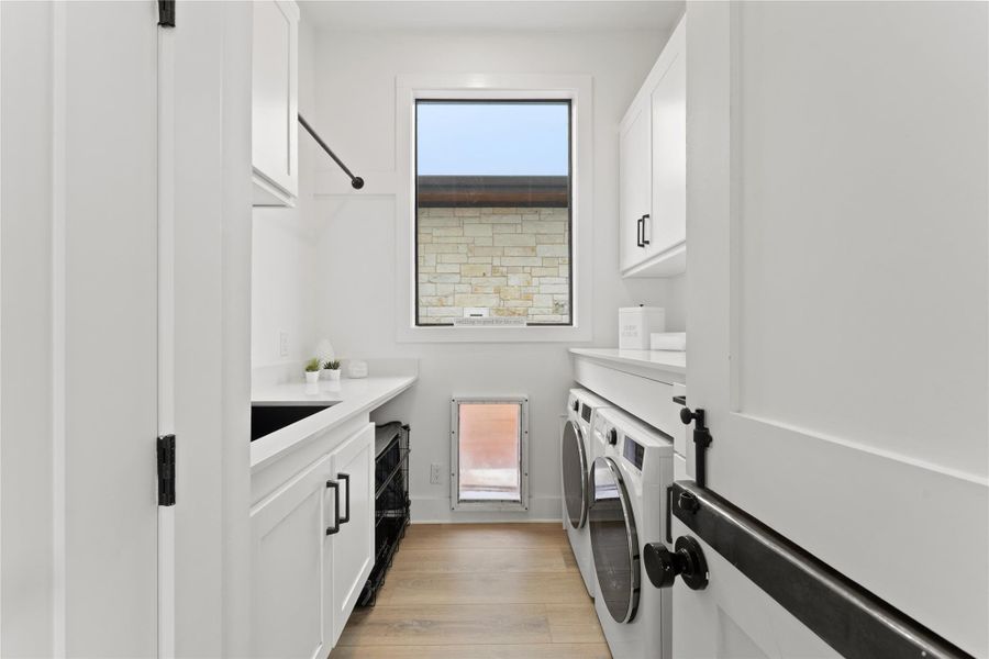 Dedicated laundry area featuring a built-in sink, white cabinetry with dark hardware, a window, and wood-finish flooring Dedicated laundry area featuring a built-in sink, white cabinetry with dark hardware, a window, and wood-finish flooring