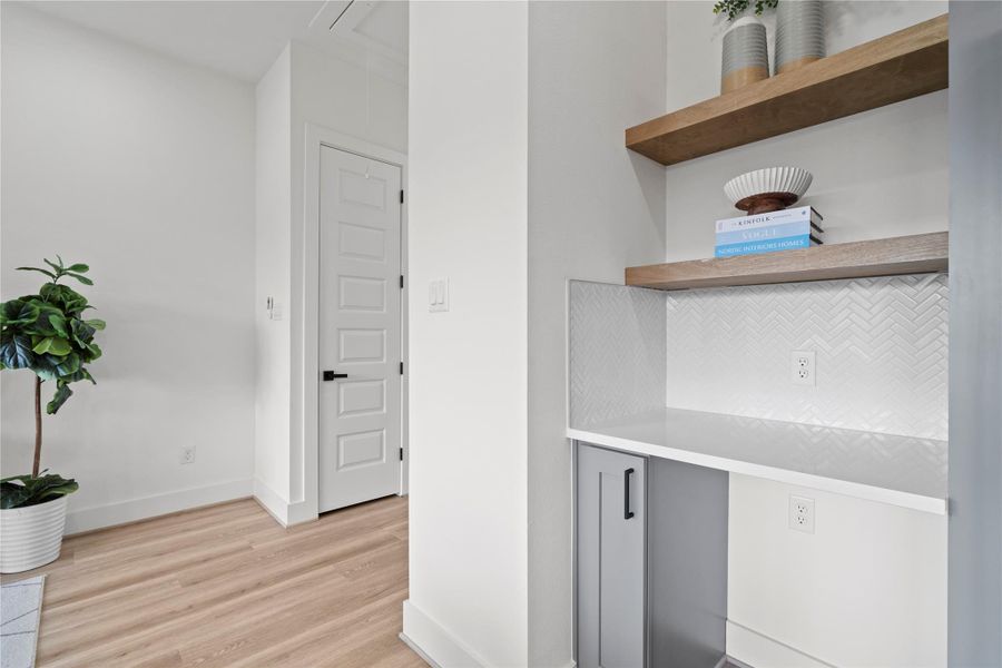 This dry bar area is designed for both style and functionality, featuring a quartz countertop, herringbone tile backsplash, and floating wood shelves for display. A dedicated space for a wine fridge adds convenience, while lower cabinetry provides concealed storage for bar essentials.