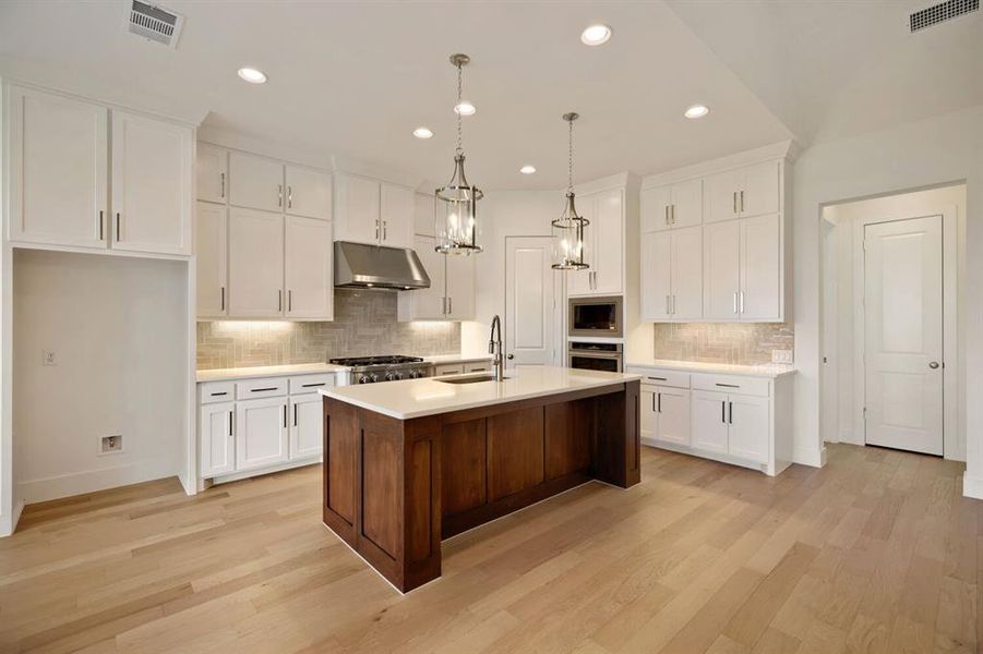 Dual tone kitchen with tasteful backsplash, light wood-type flooring, a kitchen island with sink, decorative light fixtures, and two tone color scheme