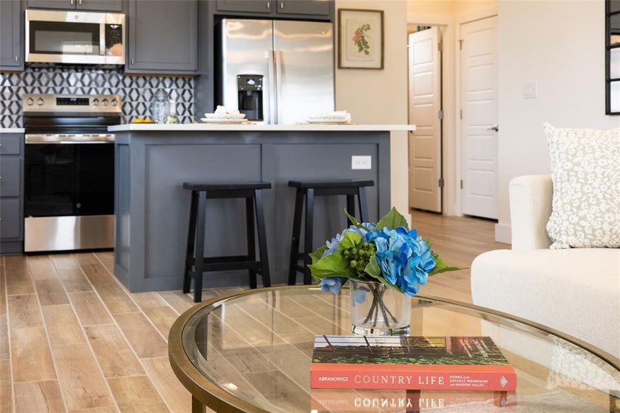 Open-concept kitchen featuring gray cabinetry, stainless steel appliances, a pattern tile backsplash, an island with countertop seating, and wood-finish flooring