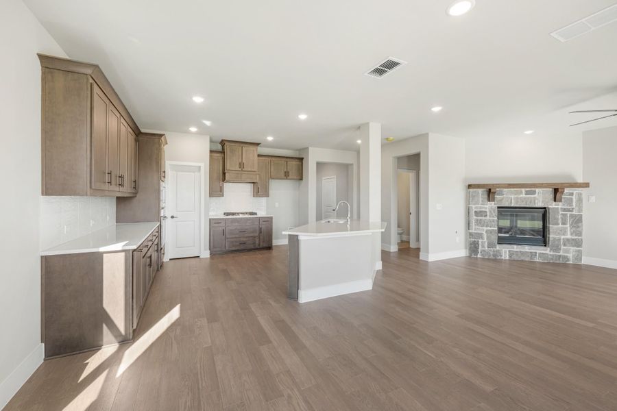 Open-concept kitchen and living area with brown cabinets, white island, hardwood floors, and stone fireplace.