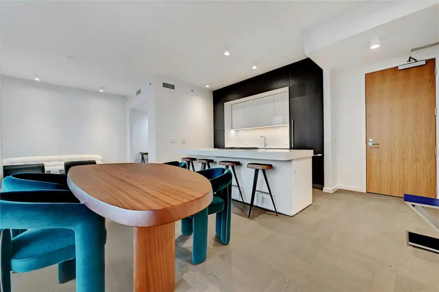 A modern, open-concept kitchen, dining and living area. The kitchen features a sleek, minimalist design with a large island at its center, topped with white-colored stone countertop. The island includes a built-in gas cooktop and stainless steel oven. Furnishings provided by Scott + Cooner available for purchase separately. Photo is model unit #2406.