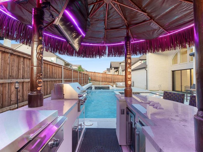 Outdoor kitchen featuring built-in stainless steel appliances, stone countertops, and a tiki-style thatched roof with perimeter lighting Outdoor kitchen featuring built-in stainless steel appliances, stone countertops, and a tiki-style thatched roof with perimeter lighting
