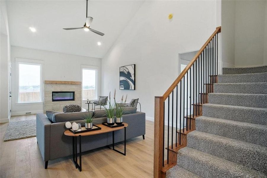 Living area with a glass covered fireplace, high vaulted ceiling, light wood-type flooring, ceiling fan, and stairs