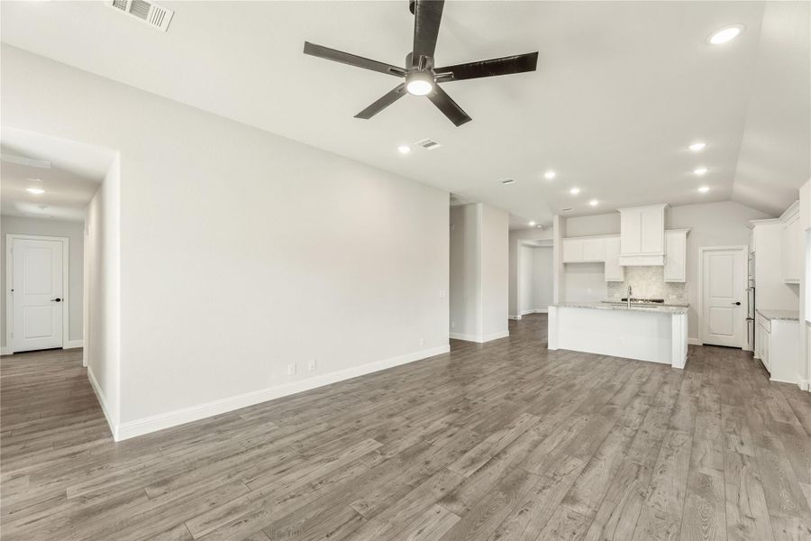 Open-concept living area with wood-look floors, ceiling fan, and white kitchen with island in background.