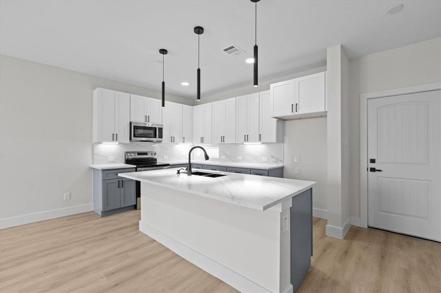 Kitchen with stainless steel appliances, light wood-style floors, hanging light fixtures, an island with sink, and white cabinets