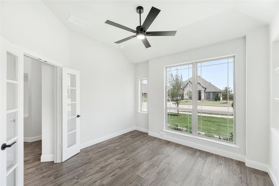 Empty room with hardwood floors, vaulted ceiling, black ceiling fan, and large windows with neighborhood view