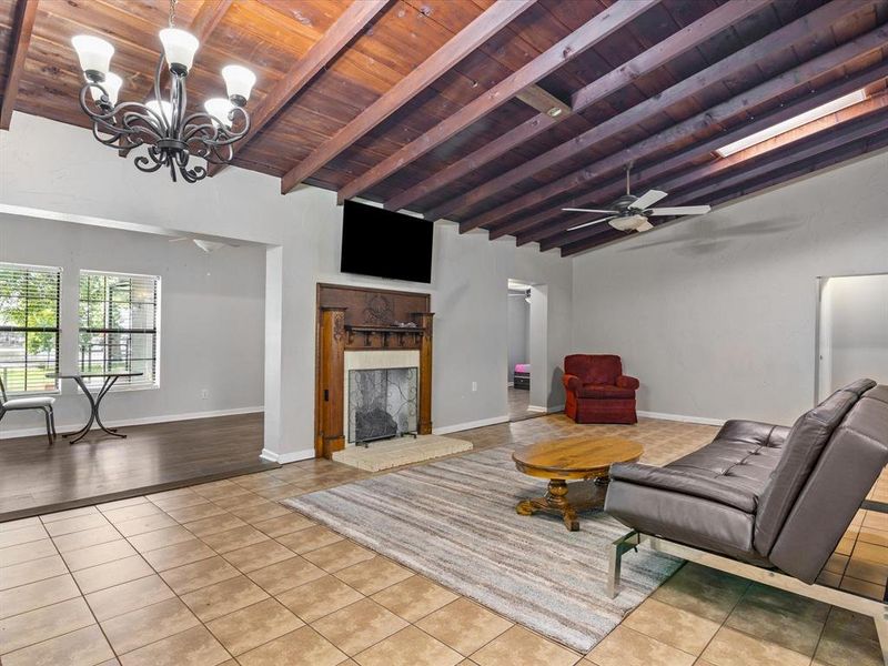 Living area with a wood ceiling with exposed beams, tile patterned floors, a ceiling fan, a chandelier, and a fireplace with raised hearth Living area with a wood ceiling with exposed beams, tile patterned floors, a ceiling fan, a chandelier, and a fireplace with raised hearth