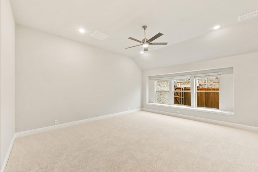 Empty bedroom with vaulted ceiling, ceiling fan, carpet flooring, and large window overlooking backyard fence Empty bedroom with vaulted ceiling, ceiling fan, carpet flooring, and large window overlooking backyard fence