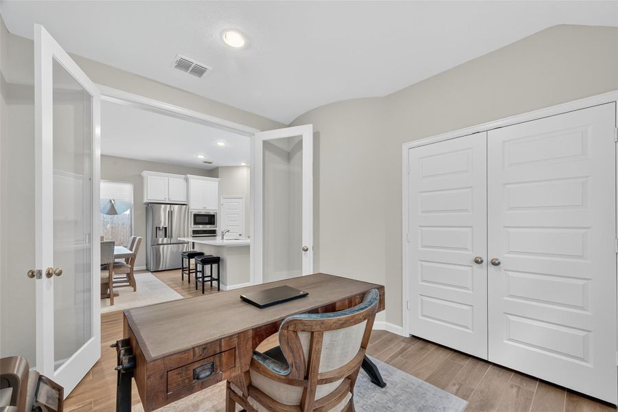 This photo showcases a bright and open home office with French doors leading to a modern kitchen and dining area. The room features a wooden desk, ample closet space, and light-colored walls, creating a cozy yet functional workspace. This photo showcases a bright and open home office with French doors leading to a modern kitchen and dining area. The room features a wooden desk, ample closet space, and light-colored walls, creating a cozy yet functional workspace.