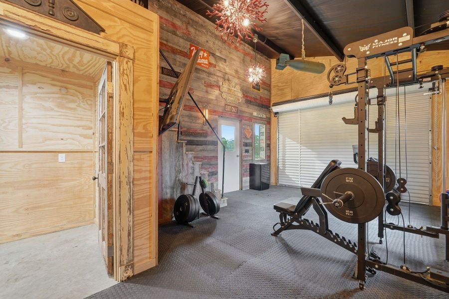 This photo showcases a home gym with exercise equipment on rubber flooring, wood-paneled walls, and a rustic aesthetic. It features a large garage door for natural light and ventilation, creating an ideal workout space.