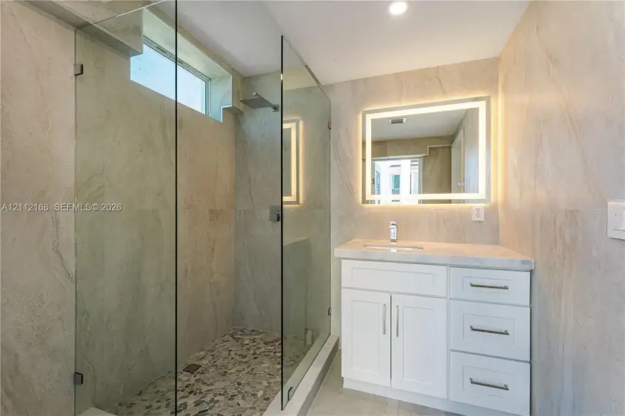 Walk-in shower with a glass enclosure, pebble tile floor, and rainfall showerhead, complemented by a white vanity with a stone countertop, integrated sink, and an illuminated mirror Walk-in shower with a glass enclosure, pebble tile floor, and rainfall showerhead, complemented by a white vanity with a stone countertop, integrated sink, and an illuminated mirror