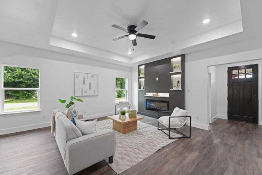 Spacious living area featuring tray ceilings with recessed lighting, a modern built-in fireplace with integrated shelving, dark wood-finish flooring, and a multi-paneled entry door Spacious living area featuring tray ceilings with recessed lighting, a modern built-in fireplace with integrated shelving, dark wood-finish flooring, and a multi-paneled entry door