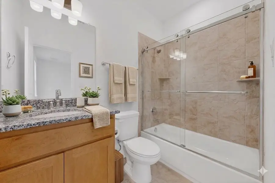 Bathroom featuring a light wood-finish vanity with a speckled countertop, a large framed mirror, and a built-in tub with sliding glass doors and tiled surround