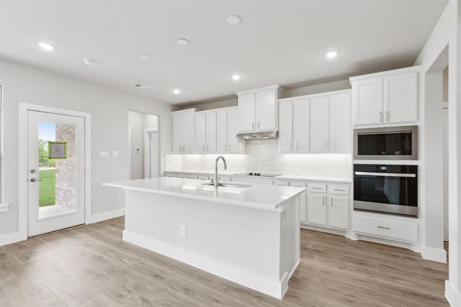 Kitchen featuring stainless steel appliances, under cabinet range hood, decorative backsplash, an island with sink, and light countertops Kitchen featuring stainless steel appliances, under cabinet range hood, decorative backsplash, an island with sink, and light countertops