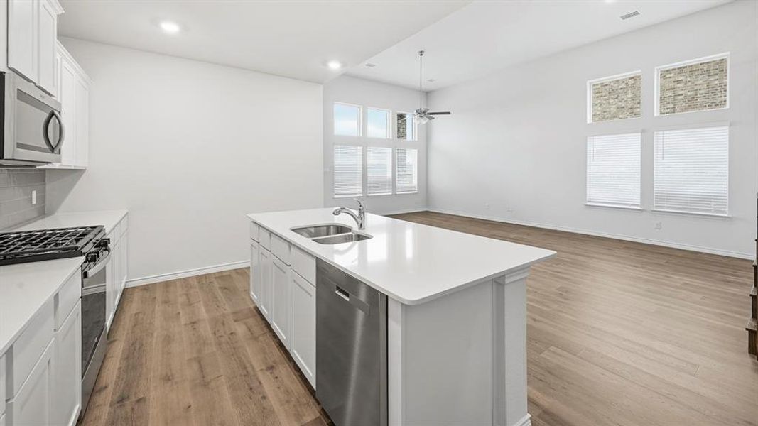 Open-concept kitchen and living area featuring white cabinetry, stainless steel appliances, a kitchen island with dual sinks, wood-finish flooring, and high ceilings with upper windows