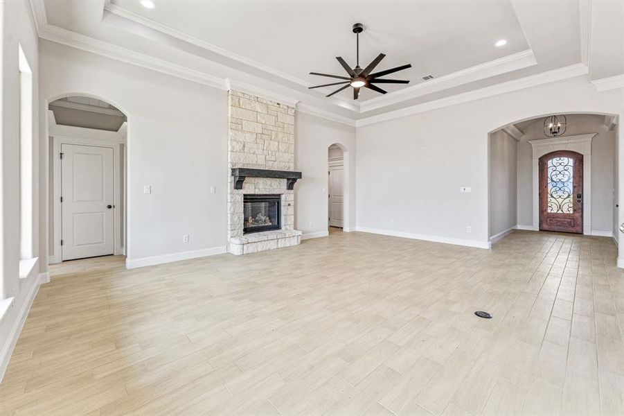 Unfurnished living room featuring arched walkways, a tray ceiling, light wood finished floors, ornamental molding, and a stone fireplace