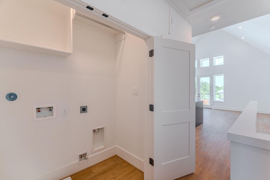 This photo shows a bright, open space with wood flooring leading into a laundry area. The room has high ceilings, large windows for natural light, and connections for appliances.Photo Disclaimer: Photos are representative of the community and may depict a similar floor plan, model home, or previously completed residence. This photo shows a bright, open space with wood flooring leading into a laundry area. The room has high ceilings, large windows for natural light, and connections for appliances.Photo Disclaimer: Photos are representative of the community and may depict a similar floor plan, model home, or previously completed residence.