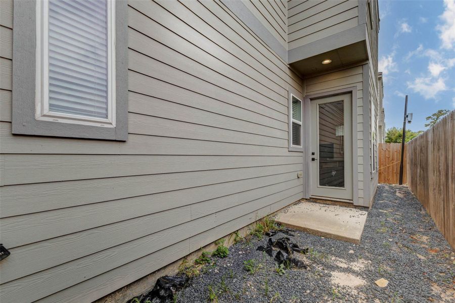 Side exterior view showing the home’s private fenced area, gravel ground cover, rear/side entry door with glass panel, exterior siding, window, and covered light. This low-maintenance outdoor space offers practical access and a defined side yard area without extensive upkeep.