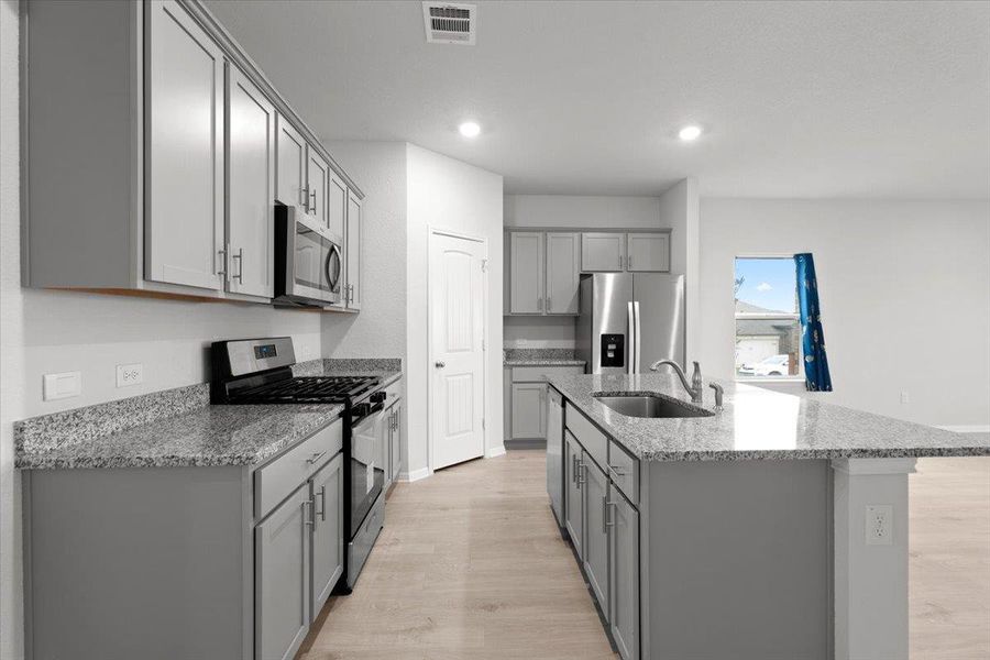 Kitchen featuring an expansive island with an undermount sink and bar seating, light gray shaker-style cabinetry, granite countertops, and stainless steel appliances, including a gas range and microwave