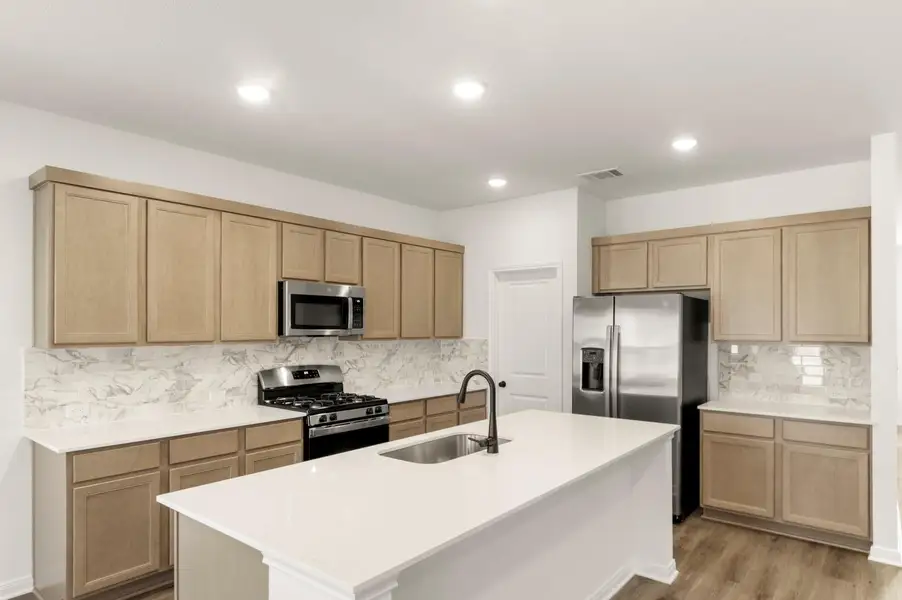 Kitchen featuring stainless steel appliances, backsplash, an island with sink, light brown cabinets, and light stone countertops Kitchen featuring stainless steel appliances, backsplash, an island with sink, light brown cabinets, and light stone countertops
