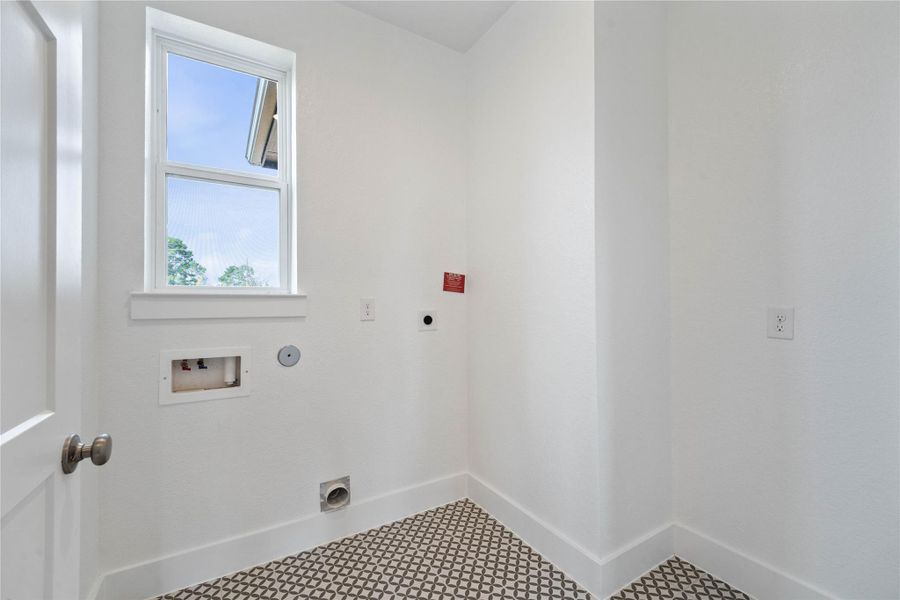 This bright and functional laundry room is conveniently located and ready for your appliances. It features clean white walls and trim, a large window providing abundant natural light, and eye-catching patterned floor tiles that add a touch of personality.