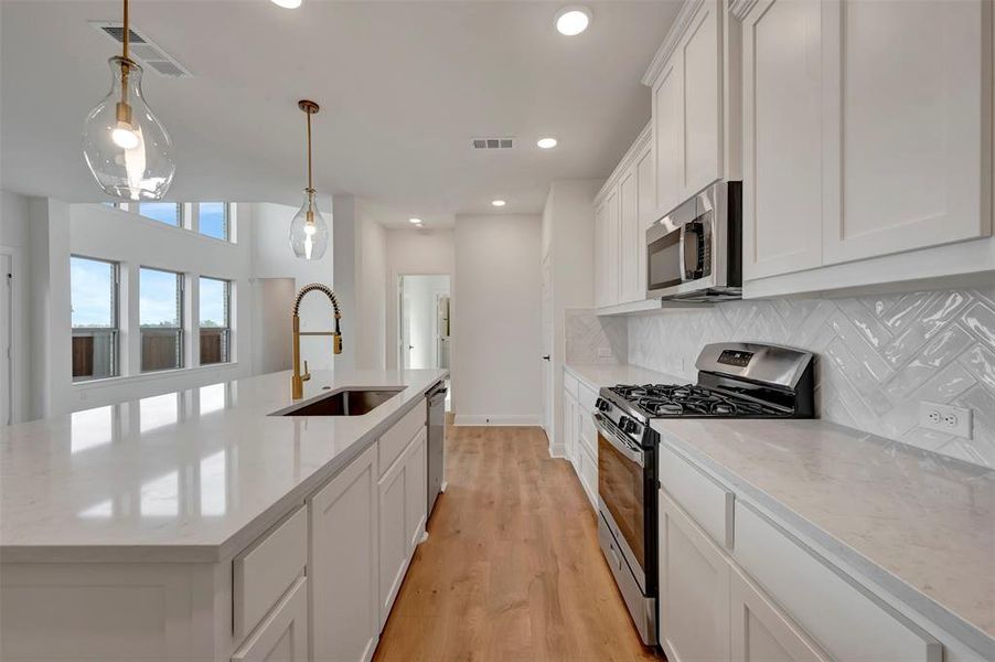 Kitchen featuring stainless steel appliances, white cabinetry, light wood finished floors, a kitchen island with sink, and hanging light fixtures