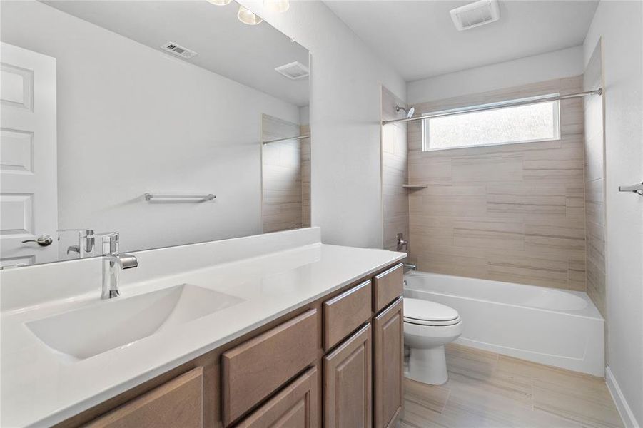 Secondary bath features tile flooring, bath/shower combo with tile surround, stained wood cabinets, beautiful light countertops, mirror, dark, sleek fixtures and modern finishes.