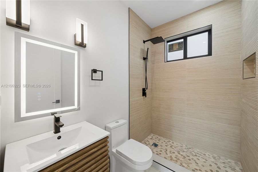 Bathroom featuring a vanity with wood-finish cabinetry, a lighted mirror, matte black wall-mounted faucet, walk-in shower with textured tile walls, and a pebble-stone floor Bathroom featuring a vanity with wood-finish cabinetry, a lighted mirror, matte black wall-mounted faucet, walk-in shower with textured tile walls, and a pebble-stone floor