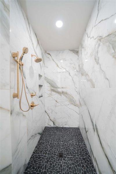 Shower enclosure featuring floor-to-ceiling veined white and grey wall tiling, a black pebble-style floor, and a gold-finish dual shower system with an integrated handheld sprayer