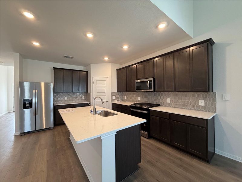 Kitchen with appliances with stainless steel finishes, dark brown cabinets, light stone countertops, tasteful backsplash, and dark wood-style floors
