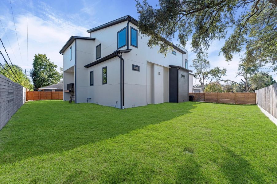 This photo shows a modern two-story home with a spacious, well-maintained backyard. The exterior features a sleek design with large windows and a mix of light and dark siding. The yard is fenced, providing privacy and ample space for outdoor activities.