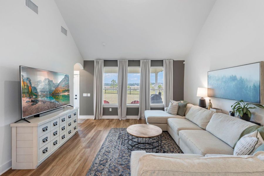 Bright living room with high ceilings, large windows, and natural light. Features a comfortable sectional, modern decor, and a flat-screen TV. Wood flooring and a tasteful rug add warmth to the space.