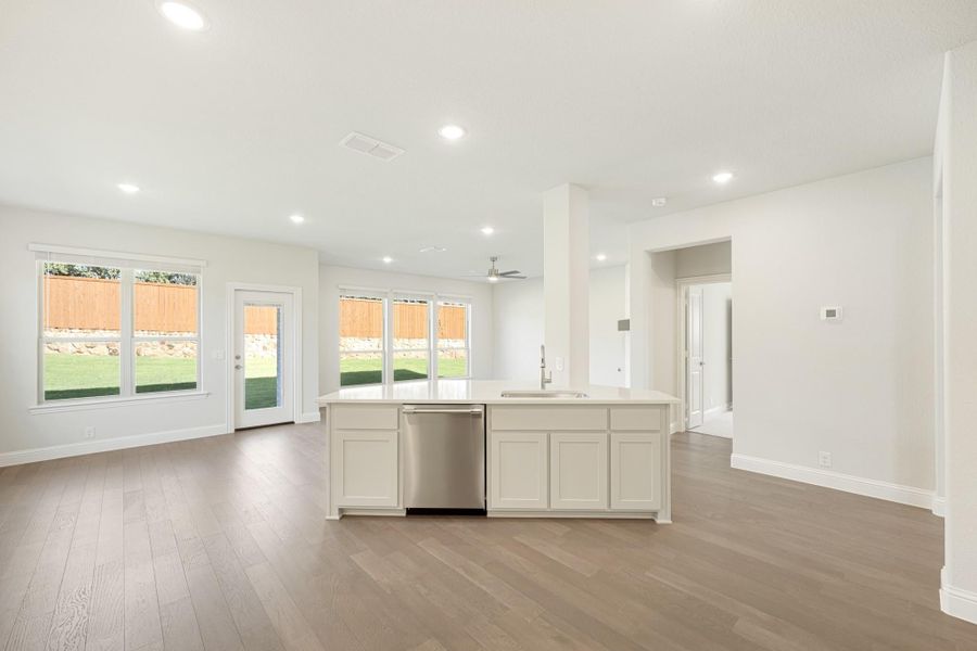 Open-concept kitchen with white island, stainless dishwasher, and light hardwood floors overlooking backyard windows