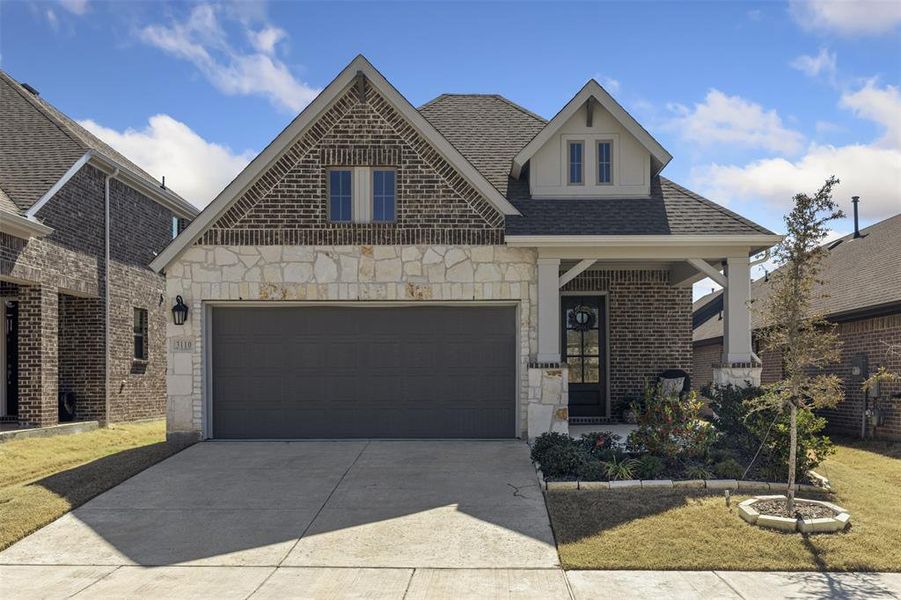 French country inspired facade featuring a garage, a shingled roof, concrete driveway, stone siding, and brick siding