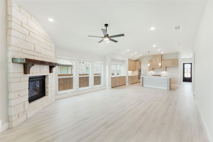 Open-concept living room with stone fireplace, ceiling fan, and light wood floors open to kitchen with island