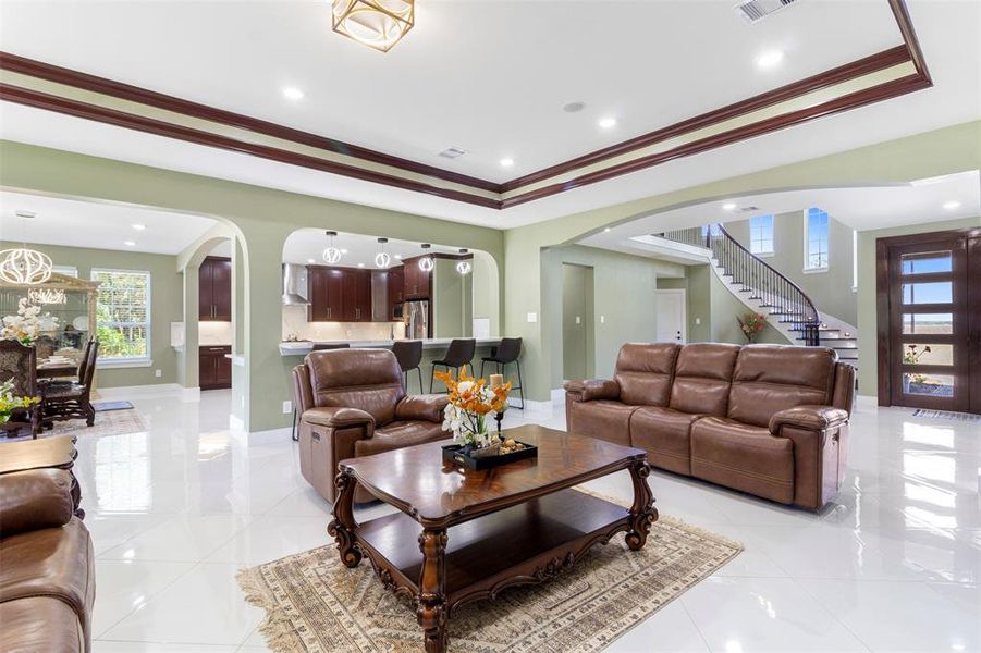 This spacious living area features elegant coffered ceilings, a modern open layout, and stylish neutral tones. It seamlessly connects to a contemporary kitchen with a breakfast bar and adjacent dining area. The room is accented by a beautiful staircase and large windows, offering ample natural light.