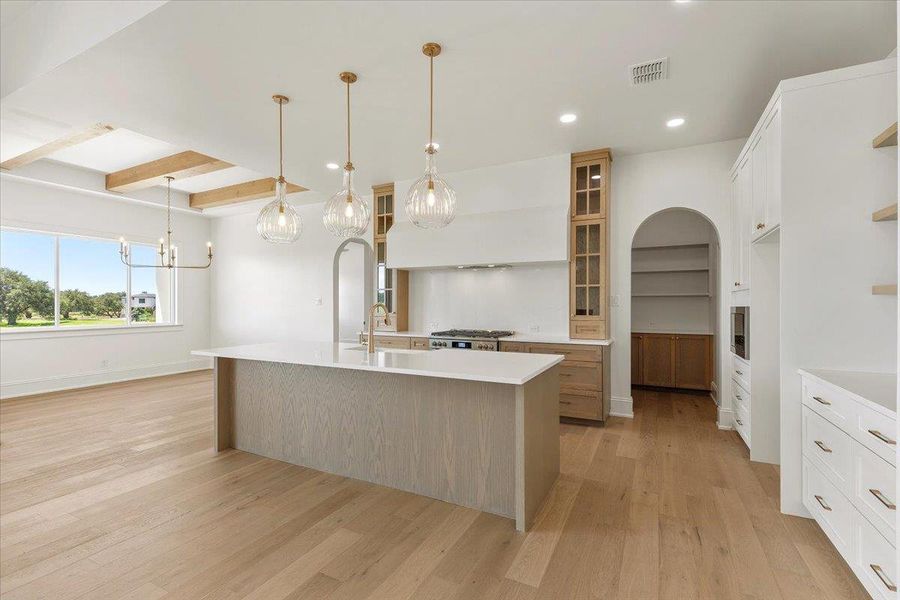 A well-lit culinary space featuring a large island, a massive custom range hood and modern overhead lighting. A well-lit culinary space featuring a large island, a massive custom range hood and modern overhead lighting.