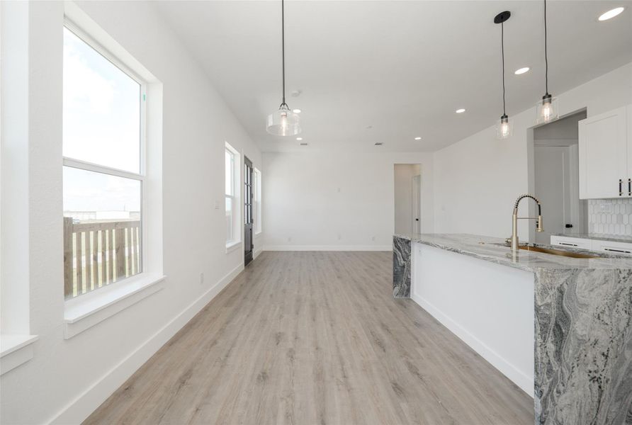 Modern kitchen with sleek white cabinets, granite countertops, and a stylish island. Features pendant lighting, stainless steel appliances, and light wood flooring. The space is bright and open, with a contemporary black door adding contrast.
