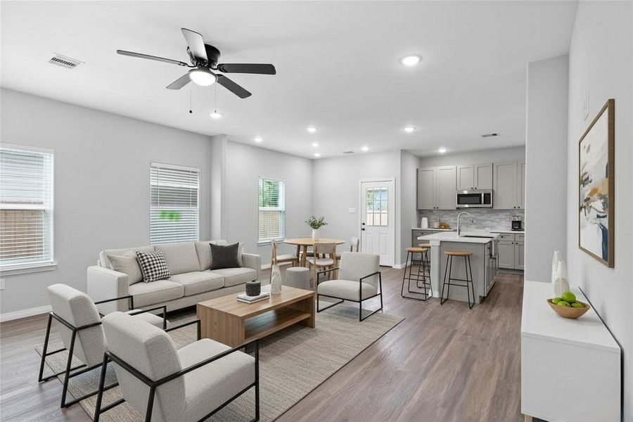 This modern, open-concept living space features a bright layout with large windows, sleek luxury vinyl plank floors, and neutral tones. The living area connects seamlessly to a stylish kitchen with a central island, stainless steel appliances, and gray cabinetry, perfect for entertaining. **Photo has been staged.**