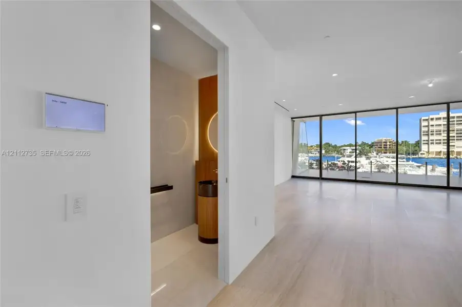 Floor-to-ceiling glass wall, wood-finish flooring, recessed lighting, and wall-mounted smart home control panel, featuring a powder room with cylindrical vanity and circular back-lit mirror