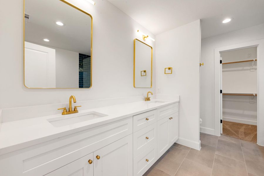 Luxurious primary bathroom featuring a double-sink vanity with quartz countertops, white cabinetry, and elegant gold fixtures. Large framed mirrors, recessed lighting, and a clean modern design create a spa-like atmosphere with direct access to the walk-in closet.