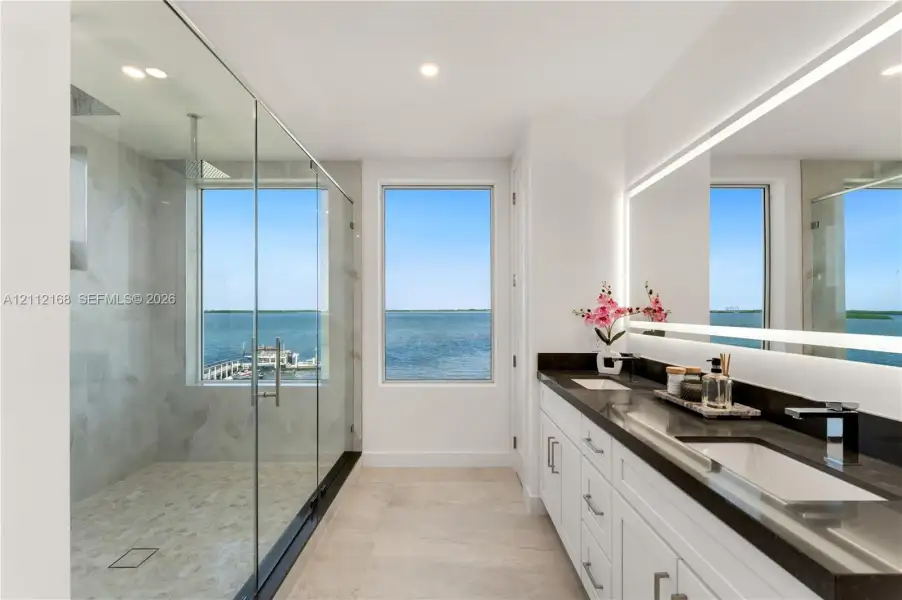 Dual-vanity bathroom featuring white cabinetry, dark stone countertops, a large frameless glass-enclosed shower with stone tile flooring, and a full-length illuminated vanity mirror Dual-vanity bathroom featuring white cabinetry, dark stone countertops, a large frameless glass-enclosed shower with stone tile flooring, and a full-length illuminated vanity mirror