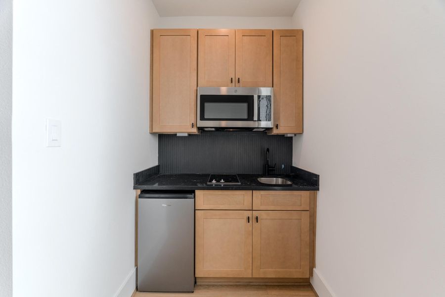 This compact kitchen features light wood cabinets, a black countertop with a small sink, a mini fridge, and a built-in microwave above the stove. It's ideal for efficient cooking in a small space.Photo Disclaimer: Photos are representative of the community and may depict a similar floor plan, model home, or previously completed residence. This compact kitchen features light wood cabinets, a black countertop with a small sink, a mini fridge, and a built-in microwave above the stove. It's ideal for efficient cooking in a small space.Photo Disclaimer: Photos are representative of the community and may depict a similar floor plan, model home, or previously completed residence.