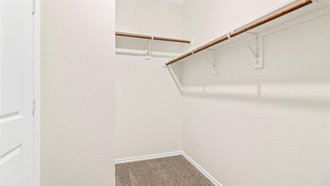 Walk-in closet featuring light beige walls, two wood-finish hanging rods with white metal brackets, and gray carpet flooring
